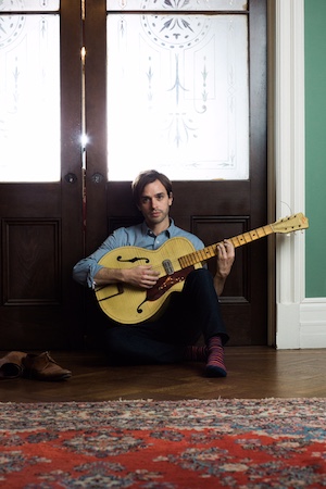 Photo of Val Emmich sitting in front of a door playing a guitar